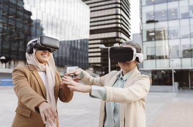 Smiling business colleagues wearing virtual reality simulator gesturing outside modern building - JCCMF05976