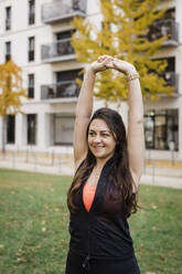 Happy woman with hands raised exercising in public park - DCRF01076