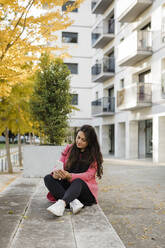 Woman with smart phone sitting on wall in front of building - DCRF01087