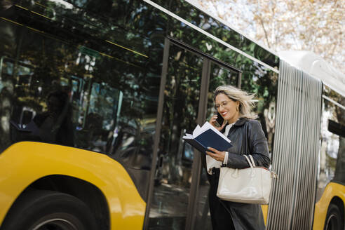 Smiling woman talking on smart phone and reading diary by bus - DCRF01106