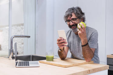 Glücklicher älterer Mann mit Apfel und Smartphone an der Kücheninsel - PNAF03458