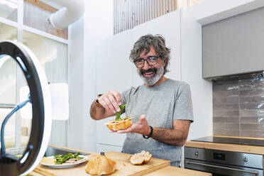 Happy senior vlogger making sandwich with croissant on kitchen island at home - PNAF03463