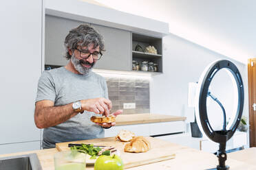 Smiling senior man making sandwich with croissant and vlogging through smart phone at home - PNAF03467