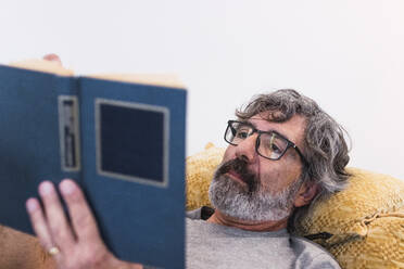 Älterer Mann liest ein Buch, während er zu Hause auf dem Sofa liegt - PNAF03471
