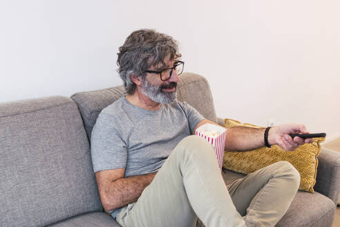 Smiling man with popcorn and remote control watching tv on sofa at home - PNAF03472