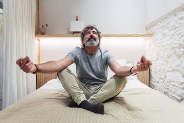 Senior man meditating on bed at home - PNAF03477