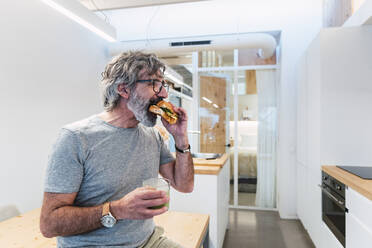 Man eating croissant sandwich with smoothie at home - PNAF03490