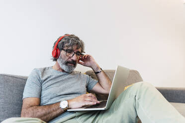 Älterer Mann sitzt auf dem Sofa, trägt Kopfhörer und benutzt zu Hause einen Laptop - PNAF03497