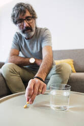 Älterer Mann mit einem Glas Wasser und einer Tablette auf dem Sofa zu Hause - PNAF03499