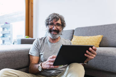 Lächelnder älterer Mann mit Tablet-PC, der zu Hause auf dem Sofa sitzt - PNAF03501