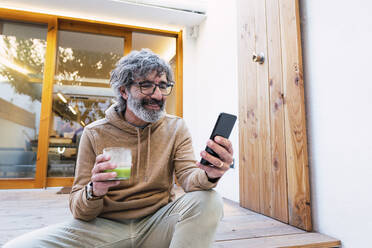 Lächelnder Mann mit einem Glas Smoothie auf der Terrasse sitzend - PNAF03503