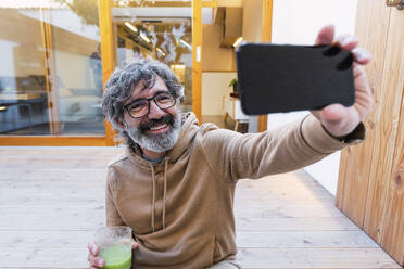 Lächelnder Mann mit einem Glas Smoothie, der ein Selfie mit seinem Mobiltelefon macht - PNAF03504