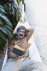 Mann beim Lesen eines Buches in der Hängematte im Hinterhof liegend - PNAF03506