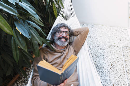 Glücklicher Mann mit Buch entspannt in Hängematte im Hinterhof - PNAF03507