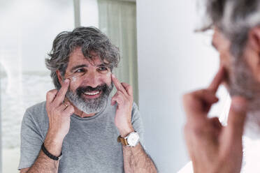 Smiling senior man looking in mirror and applying moisturizer on face - PNAF03511