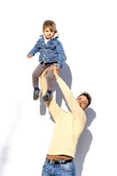 Verspielter Mann, der seinen Sohn auf die Hand nimmt, vor einer weißen Wand stehend - OCMF02364