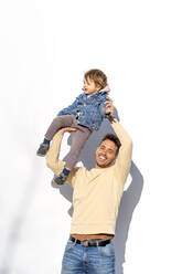 Glücklicher Mann, der seinen Sohn auf die Hand nimmt, vor einer weißen Wand stehend - OCMF02365