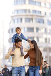 Glücklicher Mann, der seinen Sohn auf der Schulter trägt, geht mit einer jungen Frau vor einem Gebäude spazieren - OCMF02370
