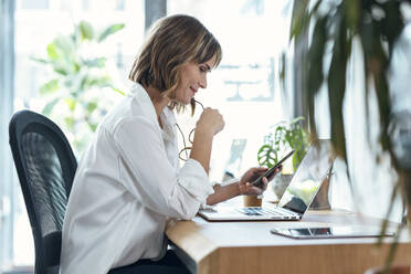 Smiling businesswoman holding eyeglasses using mobile phone at desk in office - JSRF01976