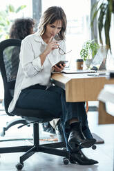 Nachdenkliche Geschäftsfrau mit Brille und Handy am Schreibtisch sitzend neben einem Kollegen im Büro - JSRF01978
