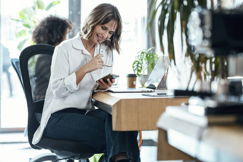 Lächelnde Geschäftsfrau, die eine Brille trägt und ein Smartphone benutzt, sitzt neben einem Kollegen am Schreibtisch im Büro - JSRF01979