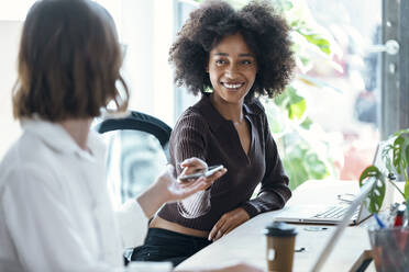 Smiling businesswoman giving mobile phone to colleague at desk in office - JSRF01984