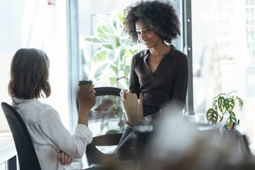 Businesswoman holding disposable cup discussing with colleague at desk in office - JSRF01989