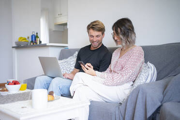 Happy freelancer with laptop looking at woman using smart phone on sofa at home - FBAF01891