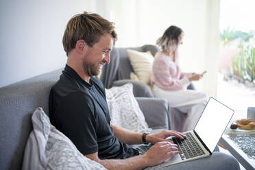 Glücklicher Freiberufler arbeitet am Laptop mit einer Frau, die ein Smartphone im Wohnzimmer benutzt - FBAF01892