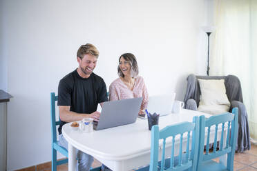 Cheerful freelancer looking at woman using laptop sitting at dining table - FBAF01905