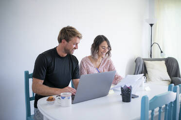 Smiling freelancer with laptop sitting by woman reading documents sitting at dining table - FBAF01909