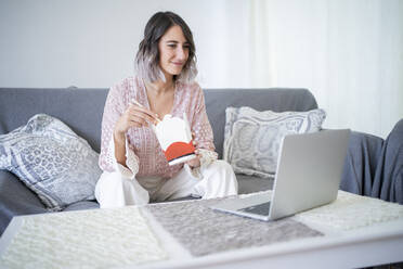 Smiling woman with take out food watching video through laptop sitting on sofa at home - FBAF01931