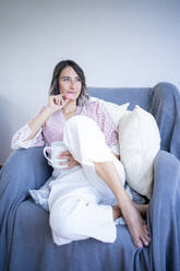 Smiling woman with macaroon and coffee mug sitting on sofa in living room - FBAF01933