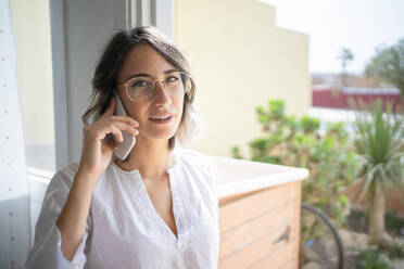 Junge Frau mit Brille, die zu Hause mit einem Mobiltelefon spricht - FBAF01938