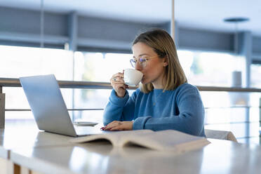 Junge Frau trinkt Kaffee und sitzt mit Laptop und Buch am Tisch - FBAF01957