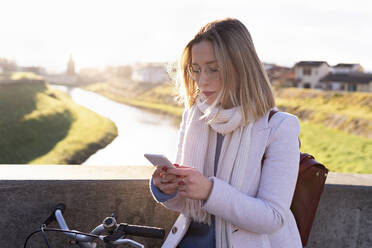 Junge Frau mit Fahrrad und Mobiltelefon an einem sonnigen Tag - FBAF01962
