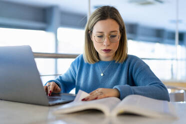 Young woman with laptop and book studying at table - FBAF01963