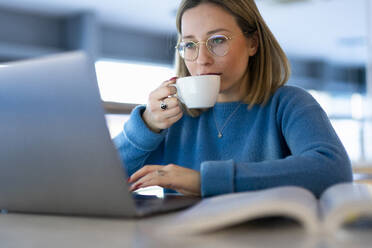 Junge Frau trinkt Kaffee und benutzt einen Laptop am Tisch - FBAF01964