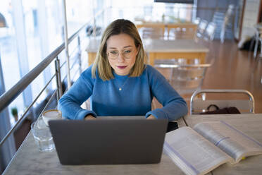 Blond young woman using laptop by book on table sitting at table - FBAF01965