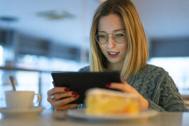 Junge Frau mit blondem Haar, die am Tisch sitzend einen Tablet-PC benutzt - FBAF01974