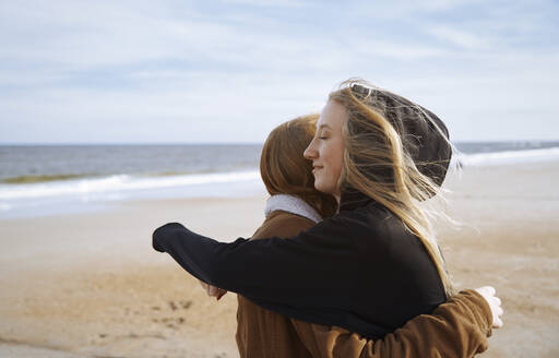 Junge Frau mit geschlossenen Augen, die ihre Schwester am Strand umarmt - AZF00393