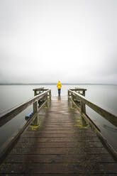 Germany, Schleswig-Holstein, Woman in yellow jacket standing on edge of lakeshore jetty - ASCF01679