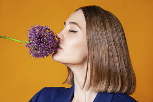 Woman with eyes closed smelling allium flower against orange background - IYNF00110
