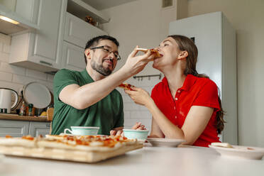 Mann füttert Frau, die am Tisch in der Küche sitzt, mit Pizza - OGF01149