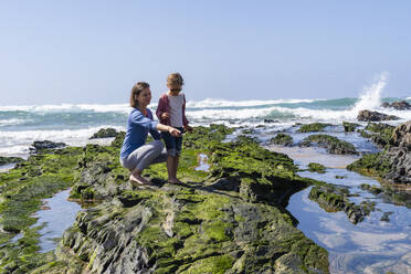 Mutter zeigt auf Tochter am felsigen Strand an einem sonnigen Tag - DIGF17786
