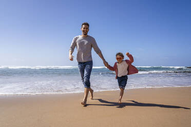 Glücklicher Vater und Tochter halten sich an den Händen und laufen am Strand an einem sonnigen Tag - DIGF17805