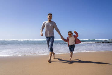 Happy father and daughter holding hands running at beach on sunny day - DIGF17805