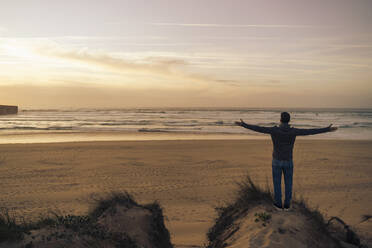 Carefree man standing on sand dune with arms outstretched at beach - DIGF17816