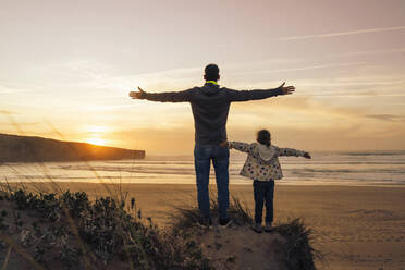 Carefree father and daughter standing on sand dune with arms outstretched at beach - DIGF17818