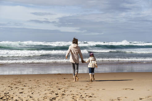 Mother and daughter walking towards waves splashing at beach on sunny day - DIGF17820